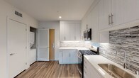 This single-floor unit showcases a contemporary design with wood-style flooring and a neutral color palette carried throughout. The open-concept living and kitchen area is bright and airy, with multiple windows allowing natural light to fill the space and a glass door leading to a private balcony with outdoor views.

The kitchen is well-appointed with floor-to-ceiling white shaker cabinetry, under-cabinet lighting, a textured mosaic tile backsplash, white quartz countertops, and a full suite of stainless steel appliances including a gas range, over-the-range microwave, refrigerator, and dishwasher. A stacked washer and dryer unit is conveniently housed within the kitchen area.

The bedrooms are generously sized with clean white walls, wood-style flooring, and ample natural light. The primary bedroom includes access to an en-suite bathroom and a walk-in closet with built-in shelving. The bathrooms are elegantly finished with marble-look porcelain tile surrounds on the soaking tubs, mosaic tile flooring, white vanities with quartz countertops, and built-in open shelving, creating a clean, spa-like aesthetic throughout.