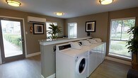 This shared laundry facility is designed with both functionality and comfort in mind. The room features a row of front-loading washers and dryers arranged along one wall, providing ample capacity for multiple users. A granite-topped counter runs alongside the machines, offering a convenient surface for sorting and folding laundry. The warm taupe walls are complemented by white trim and light wood-style flooring, creating a clean and inviting atmosphere. Natural light filters in through blinds-fitted windows, brightening the space without sacrificing privacy. Recessed and flush-mount ceiling fixtures provide consistent overhead lighting throughout. Decorative framed artwork adorns the walls, and a potted tropical plant adds a touch of greenery, elevating the room beyond a purely utilitarian space. A fire extinguisher is visibly mounted near the entrance, reflecting attention to safety standards. The overall layout is thoughtfully organized, balancing practical amenities with an aesthetic sensibility that makes routine tasks more pleasant. The single-floor design ensures easy accessibility for all users.