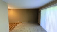 This single-floor space offers a straightforward and functional layout designed for comfortable everyday living. The main living area features wall-to-wall carpeting that extends throughout, providing a soft and warm underfoot feel, while a section of hardwood-style flooring is visible at the entry transition, adding a subtle contrast in materials. The walls are finished in a neutral earthy tone, contributing to a calm and understated ambiance throughout the space. Climate control is well-addressed, with a baseboard heating unit running along the lower portion of the main wall, supplemented by a wall-mounted air conditioning unit positioned near the window — ensuring year-round comfort. Large windows dressed with vertical blinds allow natural light to fill the room, creating a bright and airy atmosphere while also offering privacy control. The ceiling is smooth and white, helping to reflect light and enhance the sense of openness in the room. Overall, the space presents a clean, no-frills interior that prioritizes practicality and livability, making it well-suited for a range of occupants seeking a straightforward and efficiently designed living environment.