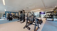 This expansive fitness center is thoughtfully designed to support a full range of workout disciplines within a single, open floor plan. The space is organized into distinct functional zones, each tailored to a specific type of training. The cardio area features a row of TRUE treadmills and elliptical machines positioned along floor-to-ceiling windows, filling the space with abundant natural light and offering views of lush greenery outside. An adjacent strength training zone houses a comprehensive selection of cable machines, weight stations, and multi-functional resistance equipment, complemented by large wall mirrors that amplify the sense of space. A dedicated group exercise and studio area provides open floor space with spin bikes, stability balls, and a storage rack for functional training accessories, making it ideal for cycling sessions or guided classes. Bold teal accent walls and architectural framing elements punctuate the otherwise neutral white and gray palette, while pendant lighting and linear LED fixtures provide consistent, well-distributed illumination throughout. Ceiling fans ensure comfortable airflow in the training areas. The flooring transitions between light-toned rubber and textured carpet tile, delineating workout zones while providing appropriate underfoot support. Overall, the facility presents a clean, professional environment well-suited for comprehensive fitness use.