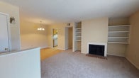 This single-floor residence offers a well-organized and comfortable layout designed for everyday living. The main living area is a standout feature, highlighted by a classic brick fireplace that serves as a natural focal point of the room. Adjacent to the fireplace, built-in shelving units provide ample display and storage space, adding both functionality and character to the room. The living area is finished with plush carpeting, contributing to a warm and inviting ambiance throughout.

A chandelier-style pendant light fixture marks the transition into the dining area, casting a soft glow that enhances the space's welcoming atmosphere. The flooring shifts to smooth hardwood in this area, creating a clear visual distinction between the living and dining zones.

The kitchen is accessible from the dining area and features wooden cabinetry, a double sink, and standard appliances, offering a practical and efficient workspace. The textured ceiling throughout the space adds a subtle architectural detail that is consistent across all rooms.

Overall, the space presents a traditional residential design with neutral tones, a logical flow between rooms, and a combination of carpet and hardwood flooring that defines each area clearly.