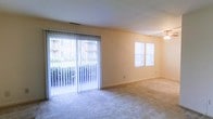 This single-floor space features a spacious, open living area finished with wall-to-wall carpeting in a neutral tone, contributing to a clean and comfortable atmosphere. The room is notably well-lit, benefiting from an abundance of natural light that enters through a large sliding glass door fitted with vertical blinds, which also provides direct access to an outdoor area with visible greenery beyond. A pair of windows on the adjacent wall further enhances the brightness of the interior. The walls are finished in a soft, warm beige tone, lending the space a welcoming and understated character. A ceiling fan is visible in the upper corner of the room, offering both functional ventilation and a subtle decorative element. The overall layout is open and unobstructed, allowing for flexible furniture arrangements. The combination of multiple light sources, neutral finishes, and outdoor connectivity makes this a practical and inviting living environment suited for a variety of uses.