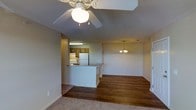 This single-floor space offers an open, flowing layout that seamlessly connects the living area to the kitchen. The living room is finished with warm, neutral carpet, while hardwood flooring extends through the adjoining dining and transitional areas, creating a natural contrast between zones. A decorative ceiling fan with a light fixture serves the living space, providing both airflow and ambient lighting.

The kitchen is positioned behind a white pass-through counter, offering a half-wall separation that maintains an open feel while defining the cooking area. Inside the kitchen, medium-toned wood cabinetry lines the walls, complemented by a white refrigerator and stove. Overhead fluorescent lighting illuminates the workspace efficiently.

The dining area is distinguished by a hanging chandelier with warm-toned shades, adding a touch of character to the space. Neutral-colored walls throughout contribute to a clean, cohesive aesthetic. White baseboards provide a crisp finish along the hardwood and carpeted floors. The overall ambiance is modest yet functional, with thoughtful lighting choices and a practical open-concept design suited for everyday living.
