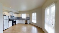This single-floor space showcases an open-concept design that seamlessly integrates the kitchen and living area into one cohesive environment. The kitchen is outfitted with crisp white cabinetry, providing ample storage across both upper and lower cabinets. A classic gray subway tile backsplash runs along the walls, adding a timeless, polished detail. Stainless steel appliances, including a gas range and dishwasher, complement the functional layout, while a single-basin sink sits beneath a window, offering a pleasant view while working in the kitchen. Light wood-toned flooring extends throughout the entire space, contributing to a warm and inviting atmosphere. Multiple windows dressed with white horizontal blinds allow generous natural light to fill the room, enhancing the open and airy feel. The living area benefits from the connected layout, offering flexible use of the space. Smooth, light-toned walls and recessed lighting further contribute to the bright, neutral palette. Overall, the space presents a clean, modern aesthetic well-suited for everyday living.