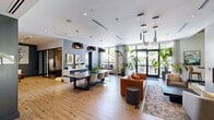 Located at 1013 Hunters Glen Dr in Plainsboro, NJ, this single-floor space offers a thoughtfully designed, open-concept layout that balances functionality with contemporary style. Light hardwood flooring extends throughout, lending warmth and continuity across the entire space.

The living area serves as the centerpiece, anchored by a striking floor-to-ceiling concrete-clad column housing a linear gas fireplace and a wall-mounted television — a bold architectural feature that draws the eye and defines the room. Plush seating arrangements in cognac leather and soft gray tones surround the fireplace, creating an inviting lounge environment.

Floor-to-ceiling grid-pane windows flood the interior with abundant natural light and provide panoramic views of the lush, landscaped outdoor courtyard, complete with lounge seating and shade umbrellas. A dedicated dining and work zone features a long communal table with modern seating beneath dramatic pendant lighting.

A built-in banquette with textured upholstered panels and octagonal mirror accents adds a distinctive, design-forward nook. Open shelving in deep navy tones provides both display and storage character. The entry foyer is framed by oversized tropical plants and glass double doors, setting an inviting tone. Recessed lighting throughout ensures a well-lit, polished ambiance at all times.