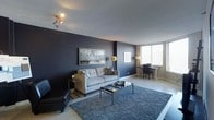 This thoughtfully designed single-floor space makes a striking first impression with its bold, contemporary aesthetic. The living area serves as the centerpiece of the space, featuring a dramatic dark navy accent wall that creates a sense of depth and sophistication. A plush grey sectional sofa and a coordinating deep-pile area rug anchor the room, lending warmth and texture to the modern palette. A sleek glass-top coffee table and curated decorative accessories complete the living arrangement with understated elegance. Track lighting on the textured ceiling adds a functional yet stylish touch, while a standing floor lamp in a gold finish introduces a refined contrast against the dark walls. A framed nature-inspired artwork adorns the accent wall, adding visual interest and a subtle organic element to the design. Adjacent to the living area, a compact dining nook is positioned to take full advantage of the large windows fitted with horizontal blinds, allowing for abundant natural light to fill the space throughout the day. The overall ambiance is modern, moody, and cohesive, blending dark and neutral tones seamlessly to create an inviting yet sophisticated atmosphere that is both functional and visually compelling.