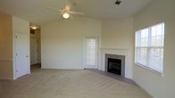 This single-story space showcases a spacious main living area defined by its vaulted ceiling, which lends a sense of openness and architectural interest. A ceiling fan with a light fixture is centrally mounted, providing both airflow and ambient lighting. The living area is finished with neutral wall-to-wall carpeting that complements the light, beige-toned walls throughout.

A tile-surround fireplace serves as a focal point along one wall, featuring a classic white mantel that adds a traditional touch to the room. Natural light filters in through a door with a windowed panel and what appears to be an additional window on the adjacent wall, keeping the interior bright and airy. The door provides direct access to what may be an outdoor area.

The overall layout is open and unobstructed, offering flexibility for various furniture arrangements. The neutral palette of carpet, walls, and trim creates a clean, cohesive aesthetic. The combination of the vaulted ceiling, fireplace, and natural light gives the space a welcoming and comfortable ambiance suitable for everyday living.