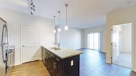 This single-floor space showcases a thoughtfully designed open-concept layout that seamlessly connects the kitchen and living areas. The kitchen is a standout feature, centered around a large island topped with speckled granite countertops and equipped with a stainless steel double sink. Dark espresso cabinetry with brushed nickel hardware provides a striking contrast against the light-toned walls. Three pendant lights hang above the island, offering both functional task lighting and a modern aesthetic touch, complemented by a track lighting fixture overhead. The flooring throughout is hardwood-style, adding warmth and continuity across the space. The open living area benefits from an abundance of natural light, primarily sourced from a full-width sliding glass door that leads to a private balcony, offering a seamless indoor-outdoor connection. The clean white walls and neutral palette create a bright, airy ambiance throughout. A paneled white door near the kitchen provides access to an additional room or storage space. The overall design balances contemporary finishes with a practical, livable layout ideal for everyday comfort.