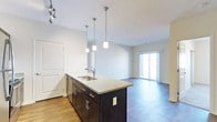 This single-floor space showcases a thoughtfully designed open-concept layout that seamlessly connects the kitchen and living areas. The kitchen is a standout feature, centered around a large island topped with speckled granite countertops and equipped with a stainless steel double sink. Dark espresso cabinetry with brushed nickel hardware provides a striking contrast against the light-toned walls. Three pendant lights hang above the island, offering both functional task lighting and a modern aesthetic touch, complemented by a track lighting fixture overhead. The flooring throughout is hardwood-style, adding warmth and continuity across the space. The open living area benefits from an abundance of natural light, primarily sourced from a full-width sliding glass door that leads to a private balcony, offering a seamless indoor-outdoor connection. The clean white walls and neutral palette create a bright, airy ambiance throughout. A paneled white door near the kitchen provides access to an additional room or storage space. The overall design balances contemporary finishes with a practical, livable layout ideal for everyday comfort.
