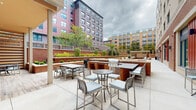 Located at 1112 First St in Alexandria, Virginia, this thoughtfully designed outdoor amenity courtyard offers a comprehensive range of recreational and social spaces. The grounds are organized into several distinct zones, each serving a specific function while contributing to a cohesive outdoor environment.

A generously sized artificial turf lawn anchors one section of the courtyard, furnished with Adirondack chairs and lawn games such as cornhole, making it ideal for casual gatherings. Adjacent to the lawn, a pergola-covered deck provides a shaded retreat with swing seating, lounge chairs, and dining furniture arranged beneath the open-air canopy.

The covered pavilion area features a warm wood-paneled ceiling, horizontal slatted privacy screens, and recessed lighting. It is equipped with comfortable lounge seating, a dining table, and a ping-pong table, offering a versatile space for both relaxation and recreation. A built-in brick fireplace within this zone adds an inviting focal point.

An open-air grilling and dining patio includes built-in gas grills framed in brick with bar-height counter seating, complemented by multiple dining tables with cushioned chairs suitable for outdoor entertaining.

Decorative brick planter beds with stone caps are arranged throughout the hardscaped areas, adding structured greenery. The surrounding architecture features classic brick facades with bold red accent doors, lending the courtyard a polished, urban character.