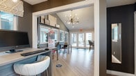 This single-floor residential leasing office and model suite presents a well-designed, contemporary space that serves both as a welcoming community hub and a showcase of available living options. The main lobby greets visitors with soaring vaulted ceilings, light hardwood flooring, and a striking sputnik-style chandelier that anchors the room with a modern aesthetic. Large French doors at the entrance flood the space with natural light and offer views of the surrounding outdoor community. Framed mirrors and a display shelf with curated décor add visual depth to the grey-toned accent walls, while branded residential signage communicates the property's management identity. A central table and seating area provide a comfortable spot for prospective residents to review information. Adjacent to the lobby, a staffed reception desk with pendant lighting and roman shades serves as the administrative hub, equipped with workstations and a TV display. A private office with glass-paneled doors is accessible from this area, offering a quieter setting for leasing consultations. The model kitchen features stainless steel appliances, including a refrigerator and range, white cabinetry with gold hardware, subway tile backsplash, open shelving, and a clean quartz countertop, demonstrating the quality of finishes available in the community's residential units.