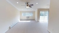 Located at 68 Mount Vernon Ave, Unit B14, in East Patchogue, NY, this single-floor residence offers a clean and comfortable layout designed for easy living. The main living area is generously proportioned, featuring plush wall-to-wall carpeting that adds warmth underfoot. Crown molding frames the ceiling, lending a refined architectural touch to the space. Recessed lighting is complemented by a ceiling fan with an integrated light fixture, providing both ambient illumination and air circulation throughout the room. Natural light pours in through a wide picture window, while a sliding glass door opens to an outdoor area, offering views of surrounding greenery and providing a seamless connection between indoor and outdoor living. Baseboard heating runs along the perimeter of the room, ensuring consistent warmth, and a wall-mounted air conditioning unit provides climate control during warmer months. The neutral, light-toned walls create a bright and airy atmosphere that serves as a versatile backdrop for a variety of interior styles. Overall, the space presents a well-maintained, functional environment with thoughtful details throughout.