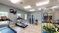 This fitness center in Foley, Alabama, offers a well-organized and fully equipped exercise environment spread across a single floor. The space is finished with light wood-look flooring and muted sage-green walls, creating a calm, focused atmosphere throughout. Bright fluorescent flush-mount lighting illuminates the entire area evenly, while multiple windows along the walls allow natural light to enter and provide views of the surrounding exterior.

The cardio section features two Spirit treadmills positioned near the windows, accompanied by a wall-mounted flat-screen television for entertainment during workouts. A True upright stationary bike and a recumbent bike offer additional low-impact cardio options, and an elliptical-style stepper rounds out the cardiovascular equipment selection.

The strength training area includes a multi-station cable machine suitable for a variety of resistance exercises, along with a dedicated weight bench placed on a protective floor mat. A rack along one wall holds medicine balls and resistance accessories, while stability balls in multiple sizes are also available for functional training.

A cork bulletin board is mounted near the glass entry doors, which open to an adjacent space. The overall layout provides ample room to move freely between equipment zones, making the fitness center practical and accessible for a range of workout routines.