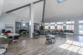 Located at 11902 Central Ave in Chino, CA, this single-story community clubhouse offers a generously sized, open-plan gathering space designed to serve a variety of social and recreational needs. The interior is defined by dramatic vaulted ceilings with exposed beam accents and skylights that flood the main hall with abundant natural light, complemented by light wood-look flooring throughout that enhances the bright, airy atmosphere.

The main gathering area features multiple dining tables and chairs arranged for group use, alongside a built-in mailbox bank and wall clock, highlighting its functional community-oriented design. A dedicated lounge zone includes comfortable seating, a wall-mounted flat-screen television with an electric fireplace unit, and curated wall art, creating a welcoming retreat atmosphere.

The full kitchen is outfitted with white cabinetry, stainless steel appliances including a refrigerator, microwave, and range, a patterned tile backsplash, and a generous light-colored countertop peninsula ideal for catering and events. Transom windows and large standard windows line the walls throughout, providing views of the surrounding landscape and pool area. A set of glass double doors opens directly to the outdoor pool area, seamlessly connecting indoor and outdoor living. Wall sconces provide warm accent lighting throughout the space.