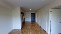 This single-floor space features a clean, modern design with warm hardwood-style flooring that runs throughout the open layout, creating a cohesive and inviting atmosphere. The open-concept arrangement connects the kitchen area to the main living space, allowing for a practical and efficient flow between functional zones.

The kitchen is positioned along one wall and is equipped with stainless steel appliances, including a refrigerator and range, complemented by dark cabinetry that provides a contrast against the light-toned walls. Recessed lighting in the ceiling offers focused illumination throughout the space.

Toward the rear of the unit, a dark-painted door serves as a visual focal point and leads to an adjacent area, while two white-paneled doors along the right wall offer access to additional rooms or storage spaces. The white walls and neutral color palette throughout contribute to a bright, airy feel. The overall design is straightforward and functional, with modern finishes that provide a move-in ready environment suitable for a variety of living arrangements.