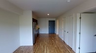 This single-floor space features a clean, modern design with warm hardwood-style flooring that runs throughout the open layout, creating a cohesive and inviting atmosphere. The open-concept arrangement connects the kitchen area to the main living space, allowing for a practical and efficient flow between functional zones.

The kitchen is positioned along one wall and is equipped with stainless steel appliances, including a refrigerator and range, complemented by dark cabinetry that provides a contrast against the light-toned walls. Recessed lighting in the ceiling offers focused illumination throughout the space.

Toward the rear of the unit, a dark-painted door serves as a visual focal point and leads to an adjacent area, while two white-paneled doors along the right wall offer access to additional rooms or storage spaces. The white walls and neutral color palette throughout contribute to a bright, airy feel. The overall design is straightforward and functional, with modern finishes that provide a move-in ready environment suitable for a variety of living arrangements.