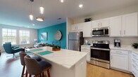 This thoughtfully designed single-floor residence showcases a cohesive, contemporary aesthetic highlighted by bold teal accent walls that appear throughout, creating a vibrant yet harmonious atmosphere across the space. The open-concept kitchen is a standout feature, outfitted with crisp white shaker cabinetry, quartz countertops, a full suite of stainless steel appliances, and a central island with bar seating illuminated by sleek pendant lighting. Light wood-look flooring flows through the kitchen and living areas, enhancing the bright, airy feel. A dedicated dining area sits adjacent to the kitchen, offering a seamless transition for entertaining. The living room is generously proportioned, featuring large double-hung windows that invite an abundance of natural light and views of the surrounding landscape. The bedroom continues the teal accent theme, with plush carpeting underfoot, dual windows for natural light, and ample room for a full suite of furnishings. A second bedroom provides additional flexibility. The bathroom features a wide vanity with white cabinetry, quartz countertops, and wood-look flooring. A laundry area with stacked washer and dryer adds everyday convenience. Overall, the space presents a polished, move-in-ready environment with a modern color palette and functional layout.