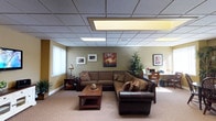 This single-floor space is thoughtfully designed to create a warm and professional atmosphere. The main lounge area serves as the centerpiece, featuring a large L-shaped sectional sofa in rich brown tones complemented by a dark leather accent piece, arranged around a solid wood coffee table. The neutral carpeting and olive-toned walls contribute to a calm, welcoming ambiance throughout.

The drop-tile ceiling is fitted with recessed panel lighting that provides consistent, even illumination across the space. A window along one wall admits natural light, softened by white vertical blinds. Decorative artwork, including a vibrant tree-themed canvas painting, adds visual interest and color to the walls.

A potted tree and additional greenery bring natural elements indoors, enhancing the inviting feel of the environment. Toward one side of the space, a small work or dining area is visible, furnished with wooden chairs and a desk, suggesting a versatile, multi-purpose layout. A side table with a lamp and a small bookshelf round out the lounge area, offering both practicality and warmth.

Overall, this space strikes a balance between comfort and functionality, making it well-suited for use as a professional office, counseling suite, or client-facing reception and waiting area.