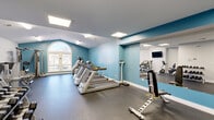 Located at 1080 Valley Bluff Dr in Perrysburg, Ohio, this well-equipped fitness center offers a comprehensive workout environment within a single, open-plan room. The space is defined by a vaulted ceiling that lends an airy, spacious feel, complemented by a large arched window that invites natural light and outdoor views. Teal accent walls contrast with crisp white surfaces, creating a clean and energizing atmosphere throughout.

The cardio zone is anchored by a row of Matrix treadmills and elliptical trainers, ideal for sustained aerobic workouts. Strength training options include Hoist cable and resistance machines positioned along one side of the room, alongside a multi-function weight bench and a dedicated dumbbell rack stocked with a full range of free weights. A wall-mounted flat-screen television adds an entertainment element to both the cardio and strength areas.

A large floor-to-ceiling mirror spans one wall, visually expanding the space and allowing users to monitor their form during exercise. The durable rubber flooring provides both safety and comfort underfoot. Recessed and flush-mounted panel lighting ensures the space remains well-lit throughout. The fitness center also includes a wall-mounted scale and a small bench for added convenience, making it a fully functional facility for a variety of fitness routines.