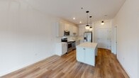 Located at 301 N La Fox St in South Elgin, Illinois, this single-floor residence offers a thoughtfully designed layout with modern finishes throughout. The kitchen serves as a centerpiece of the home, showcasing white shaker-style cabinetry extending to the ceiling, a classic subway tile backsplash, and a full suite of stainless steel appliances including a range, microwave, dishwasher, and refrigerator. A central island with a clean white countertop provides additional prep space and casual seating, while globe pendant lights add a contemporary touch. Warm-toned hardwood-style flooring flows throughout the kitchen and main living areas, creating a cohesive and inviting atmosphere. The bedroom areas feature plush neutral carpeting and crisp white walls, with a generously sized window allowing natural light to brighten the space. A hallway connects the living areas to the bedrooms and bathroom, maintaining an efficient and open flow. The bathroom is finished with hardwood-style flooring, a white vanity, and a tiled walk-in shower enclosed by a sliding glass door with chrome hardware. The overall design aesthetic is clean and contemporary, with a neutral palette that offers a bright, move-in-ready environment suited for comfortable everyday living.