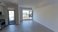 This contemporary single-floor unit offers a clean, open-concept design that maximizes both space and natural light. The living area is anchored by large floor-to-ceiling windows with sleek dark frames, flooding the interior with daylight and providing sweeping views of the surrounding urban cityscape. The light-toned hardwood flooring runs seamlessly throughout, contributing to a cohesive and airy aesthetic.

The kitchen is positioned along one wall and features white cabinetry and stainless steel appliances, including a range, integrating efficiently into the open-plan layout without disrupting the flow of the living space. A private balcony is accessible through a glass-panel door adjacent to the kitchen, offering an outdoor retreat with additional city views.

The interior walls are finished in crisp white, complementing the minimalist design and enhancing the sense of openness. Recessed track lighting on the ceiling provides functional illumination throughout. The overall ambiance is modern and understated, well-suited for those who appreciate clean lines, abundant natural light, and an urban living environment. The layout is versatile, allowing for flexible furniture arrangements within the main living and dining areas.