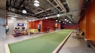 The Community Room at 121 Westmoreland Ave in White Plains, NY is a spacious, multi-use amenity space designed to foster social interaction and recreation. The room's industrial-style aesthetic is defined by an exposed ceiling with visible ductwork, concrete columns, and pendant lighting, creating an urban, loft-like atmosphere throughout.

The centerpiece of the space is a full-length bocce ball court surfaced in green turf and framed by wooden borders, flanked by benches for spectators. Bold, warm terracotta-red accent walls run along much of the room, energizing the environment and providing a striking visual contrast against concrete structural elements.

Multiple distinct seating zones are distributed throughout. One lounge area features a bright yellow sectional sofa, natural wood coffee tables, and a mounted flat-screen TV, positioned near a snack and beverage vending station. Another corner showcases a teal sectional sofa set against a sage-green accent wall, offering a more intimate gathering spot. Additional high-top tables and chairs, a pool table, and open floor space provide further options for socializing and gaming.

A large, colorful mural adorns one wall, adding an artistic focal point. A staircase with a brick surround connects the room to upper levels. The overall ambiance is lively, welcoming, and well-suited for community events and casual recreation.
