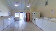 This single-floor communal laundry facility is designed for both functionality and comfort. The room is finished with light-toned tile flooring that extends throughout the space, making for easy maintenance and a clean, welcoming appearance. Overhead, white-painted exposed beam ceilings lend the room a distinctive architectural character, while two ceiling fans with light fixtures ensure consistent airflow and comfortable temperatures during use. Multiple stacked and standalone washer and dryer units line the walls, offering ample capacity for residents. A classic Coca-Cola vending machine adds a convenient touch near the entrance. Natural light streams in through a large window and an open doorway that leads to an outdoor stairway, creating a bright and pleasant environment. The open entryway also provides a view of landscaped surroundings, enhancing the overall ambiance of the space. The layout is thoughtfully arranged to allow easy movement between machines, making this laundry room both practical and comfortable for everyday use.