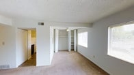 This single-story residence offers a straightforward and functional layout designed for comfortable everyday living. The main living area features neutral-toned wall-to-wall carpeting that lends warmth to the space, complemented by clean white walls and a textured ceiling. A ceiling fan provides year-round comfort and gentle air circulation throughout the room. A standout feature is the built-in open shelving unit, offering convenient storage and display space, flanked by bifold closet doors that maximize accessibility while maintaining a tidy appearance. A window along the exterior wall allows natural light to filter into the room, brightening the interior. An HVAC vent is integrated into the upper wall, ensuring efficient climate control. The layout transitions seamlessly into a tiled area that leads to the kitchen, which is partially visible and features what appears to be standard appliances. The yellow-toned walls of the adjacent area add a subtle contrast to the otherwise neutral palette of the main space. Overall, the residence presents a clean, move-in-ready environment with practical storage solutions and a cohesive, understated aesthetic throughout its single level.
