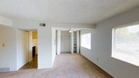 This single-story residence offers a straightforward and functional layout designed for comfortable everyday living. The main living area features neutral-toned wall-to-wall carpeting that lends warmth to the space, complemented by clean white walls and a textured ceiling. A ceiling fan provides year-round comfort and gentle air circulation throughout the room. A standout feature is the built-in open shelving unit, offering convenient storage and display space, flanked by bifold closet doors that maximize accessibility while maintaining a tidy appearance. A window along the exterior wall allows natural light to filter into the room, brightening the interior. An HVAC vent is integrated into the upper wall, ensuring efficient climate control. The layout transitions seamlessly into a tiled area that leads to the kitchen, which is partially visible and features what appears to be standard appliances. The yellow-toned walls of the adjacent area add a subtle contrast to the otherwise neutral palette of the main space. Overall, the residence presents a clean, move-in-ready environment with practical storage solutions and a cohesive, understated aesthetic throughout its single level.