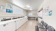 This shared laundry facility is a thoughtfully organized, single-room space designed to accommodate multiple users efficiently. The interior features light wood-look flooring, neutral beige walls, and a textured ceiling fitted with flush-mount lighting fixtures that provide consistent, even illumination throughout. Natural light filters in through multiple windows fitted with white horizontal blinds, enhancing the bright and clean atmosphere of the room.

Along one wall, a long row of white top-load washing machines is neatly arranged side by side, complemented by a front-load washer at the end. On the opposite side, stacked dryer units are built into an alcove, each equipped with individual coin-operated or card-reader controls. A continuous countertop folding station runs along one wall, supported by wire laundry carts on wheels that provide convenient transport of items throughout the space. An additional wall-mounted shelf offers extra storage.

Seating is provided by a row of sturdy gray chairs positioned near the entrance, allowing users to wait comfortably while their laundry cycles complete. A bold black front entry door with glass panel inserts serves as the room's main access point. The overall layout is efficient and user-friendly, with clearly posted instructional and informational signage displayed throughout the room to guide users.