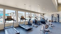 This generously sized fitness center offers a comprehensive workout environment across a single, open floor. The space is defined by soaring ceilings in the free weights area and a wall of large windows that line the perimeter, flooding the room with natural light and providing clear views of an outdoor pool and surrounding community. The durable, cushioned rubber flooring in a speckled blue-grey tone covers the entire space, providing comfort and support underfoot throughout.

The cardio section is well-appointed with multiple treadmills, elliptical trainers, stationary bikes, a rowing machine, and a stair climber, all positioned to take advantage of the outdoor views. A dedicated strength training area features a full rack of dumbbells, a cable crossover machine, and a variety of selectorized strength machines. Stability balls in multiple sizes are neatly organized on a vertical rack alongside resistance training accessories.

A decorative tropical-themed mural adds a vibrant visual accent to the strength area, while large mirrors along one wall enhance the sense of space. Ceiling fans and wall-mounted fans ensure comfortable airflow throughout. Recessed lighting supplements the natural light, and mounted flat-screen televisions provide entertainment during workouts. The space is bright, clean, and thoughtfully organized for a complete fitness experience.