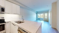 This efficiently designed one-bedroom, one-bathroom unit offers a thoughtfully organized layout across a single floor. The open-concept kitchen and living area forms the heart of the space, where a sleek kitchen island with a marble-look quartz countertop serves as both a functional workspace and casual dining spot. The kitchen is outfitted with white shaker-style cabinetry, stainless steel appliances including a refrigerator, electric range, and over-the-range microwave, along with an undermount double sink and under-cabinet lighting. Light wood-look flooring flows throughout, enhancing the bright, clean aesthetic. The living area extends toward a sliding glass door that opens to a private balcony, framing views of surrounding greenery and mountains beyond. The bedroom is a comfortable and well-lit retreat, featuring a large window with scenic mountain and treetop views, sliding glass door access to an outdoor space, and a dedicated closet equipped with built-in shelving and hanging rods for organized storage. The bathroom is finished with clean white tile surrounding a soaker tub and shower combination, complemented by modern chrome fixtures. The overall design emphasizes a contemporary, minimalist aesthetic with a neutral color palette and abundant natural light throughout.