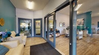 This single-floor commercial office space is thoughtfully designed to balance professional functionality with a warm, inviting atmosphere. The entry vestibule greets visitors with blue-framed glass doors, a textured entry mat, and soft overhead lighting, creating a clean and organized first impression with views into the exterior surroundings.

Beyond the entryway, the main work area opens into a spacious, airy room featuring warm wood-look flooring, vaulted ceilings, and large windows that allow generous natural light to fill the space. A prominent teal accent wall adds a bold pop of color and serves as a vibrant focal point throughout the office. Workstations with white farmhouse-style desks and modern task chairs are positioned to encourage a collaborative workflow, while shelving and storage units line the walls for practical organization.

A dedicated private office area offers a more focused workspace, complete with additional desks, white guest chairs, computer equipment, and a bold teal wall adorned with artwork — creating an environment that is both productive and visually engaging.

A cozy lounge corner provides a comfortable retreat, featuring a cream leather armchair, a patterned area rug, decorative accessories, and a stylish side table — offering a welcoming spot for informal meetings or relaxation. A built-in counter with bar seating and a computer workstation serves as a convenient resident or client resource station, complemented by large windows overlooking greenery outside.