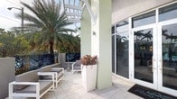 This single-story space showcases a thoughtfully designed covered outdoor patio that blends contemporary architecture with a tropical aesthetic. The patio is anchored by elegant arched columns painted in a soft sage green, which frame the space and provide a sense of structure while maintaining an open, airy feel. Overhead, a white pergola-style lattice roof allows natural light to filter through, illuminating the stone tile flooring below.

The seating area is furnished with sleek, modern outdoor furniture featuring white powder-coated frames and neutral gray cushions, arranged to encourage conversation and relaxation. A striking tall white planter serves as a focal point, housing a vibrant bromeliad with deep burgundy and green foliage. The surrounding landscape is lush with mature palm trees and tropical greenery, creating a private, resort-like atmosphere.

A decorative mosaic-tiled wall panel adds an artistic element to the background, while contemporary wall-mounted lighting ensures the space is functional after dark. Large floor-to-ceiling glass doors and windows with white frames connect the patio to the interior, allowing seamless indoor-outdoor flow and an abundance of natural light throughout.