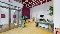 This beautifully appointed lobby makes an immediate impression with its dramatic double-height ceilings and a bold design aesthetic that blends classical and eclectic influences. The ceiling is a standout architectural feature, showcasing a deep crimson and white coffered grid pattern fitted with recessed lighting that casts a warm, inviting glow throughout the space. Beneath it, distinctive yellow Moroccan-style patterned floor tiles add a rich, tactile warmth that anchors the room's vibrant color palette.

The entry is framed by floor-to-ceiling glass windows and doors, flooding the interior with abundant natural light and offering clear views of the surrounding streetscape. Patterned deep-rose wallcovering on a feature wall adds depth and texture, complementing the crisp white wainscoting and panel molding on adjacent walls.

Furnishings are carefully curated and eclectic in character — a hand-carved, turquoise-patinated console table serves as a central focal point, adorned with floral arrangements and decorative objects. A warm caramel leather sofa, rustic wood media cabinet, and a mix of vintage and contemporary accent pieces contribute to the layered, welcoming ambiance. Lush tropical palms and oversized leafy plants are positioned throughout, bringing life and vitality to the space. A discreet reception window provides a professional service point, making this lobby both functional and visually impressive.