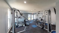 This fitness center is a thoughtfully designed, single-floor facility built to support a comprehensive workout routine. The space is finished with neutral gray walls and durable, dark modular carpet tiles accented by blue exercise mat squares, creating a clean and functional aesthetic throughout.

The gym is outfitted with a solid selection of commercial-grade equipment to accommodate both strength training and cardiovascular exercise. Strength training is well represented with multiple cable pulley machines, including an Inflight Fitness multi-station unit and additional cable systems ideal for lat pulldowns, rows, and arm exercises. A dedicated weight bench and a Cybex rowing machine round out the strength offerings.

For cardio, the facility includes at least one treadmill, positioned near a window to allow natural light into the workout space. Wall-mounted flat-screen televisions are present throughout, providing entertainment during workouts. The space also features a wall-mounted sanitizing station, promoting cleanliness and hygiene.

Natural light enters the facility through multiple windows and glass-panel doors, which also provide views of the surrounding exterior. Recessed ceiling lighting supplements the natural light for a consistently bright environment. The layout is open and well-organized, allowing users to move freely between equipment. Emergency exit signage is clearly visible, and the space is climate-controlled for year-round comfort.
