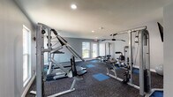 This fitness center is a thoughtfully designed, single-floor facility built to support a comprehensive workout routine. The space is finished with neutral gray walls and durable, dark modular carpet tiles accented by blue exercise mat squares, creating a clean and functional aesthetic throughout.

The gym is outfitted with a solid selection of commercial-grade equipment to accommodate both strength training and cardiovascular exercise. Strength training is well represented with multiple cable pulley machines, including an Inflight Fitness multi-station unit and additional cable systems ideal for lat pulldowns, rows, and arm exercises. A dedicated weight bench and a Cybex rowing machine round out the strength offerings.

For cardio, the facility includes at least one treadmill, positioned near a window to allow natural light into the workout space. Wall-mounted flat-screen televisions are present throughout, providing entertainment during workouts. The space also features a wall-mounted sanitizing station, promoting cleanliness and hygiene.

Natural light enters the facility through multiple windows and glass-panel doors, which also provide views of the surrounding exterior. Recessed ceiling lighting supplements the natural light for a consistently bright environment. The layout is open and well-organized, allowing users to move freely between equipment. Emergency exit signage is clearly visible, and the space is climate-controlled for year-round comfort.