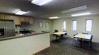 This single-floor space is designed to serve as a functional and versatile environment, blending a kitchenette with a dedicated meeting or dining area. The kitchenette is equipped with a stove, microwave, and cabinetry, making it well-suited for light meal preparation or catering during events. A dark laminate countertop defines the boundary between the kitchen zone and the open gathering area.

The main area features patterned commercial-grade carpeting and is furnished with rectangular tables and stackable chairs, accommodating multiple seating arrangements for meetings, training sessions, or group dining. The layout is open and adaptable, allowing furniture to be rearranged as needed.

Walls are finished in a neutral sage green tone, contributing to a calm, focused atmosphere. A textured ceiling hosts recessed fluorescent lighting that provides consistent, even illumination throughout the space. Natural light supplements the interior lighting through several windows fitted with blinds along one wall, offering a connection to the outdoors while maintaining privacy. Framed artwork on the walls adds a modest decorative touch. Overall, this space is practical and organized, well-suited for professional or community use.