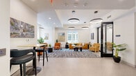 Located at 118 N Prince St in the heart of Lancaster, PA, this thoughtfully designed single-floor space offers a polished and functional environment across three distinct areas. The open-plan layout is unified by a cohesive modern aesthetic, featuring crisp white walls, light flooring, and bold circular ring pendant lighting throughout.

The living lounge serves as the centerpiece, furnished with plush burnt-orange upholstered chairs and a caramel-toned sectional sofa arranged over a large abstract patterned area rug. Large windows flood the area with natural light and provide views of the surrounding streetscape, while curated artwork and lush greenery add warmth and character.

The private meeting room offers an intimate setting anchored by a sleek, organic-shaped wooden conference table surrounded by textured golden accent chairs. A circular LED ceiling fixture and a colorful geometric art piece on the wall contribute to a creative yet professional atmosphere.

The entry and lobby area features built-in shelving with colorful decorative objects, cabinetry in a muted sage tone, and a high-top bar seating nook — ideal for casual work or informal conversation. The space flows seamlessly between zones, making it well-suited for both collaborative gatherings and focused work sessions.