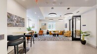 Located at 118 N Prince St in the heart of Lancaster, PA, this thoughtfully designed single-floor space offers a polished and functional environment across three distinct areas. The open-plan layout is unified by a cohesive modern aesthetic, featuring crisp white walls, light flooring, and bold circular ring pendant lighting throughout.

The living lounge serves as the centerpiece, furnished with plush burnt-orange upholstered chairs and a caramel-toned sectional sofa arranged over a large abstract patterned area rug. Large windows flood the area with natural light and provide views of the surrounding streetscape, while curated artwork and lush greenery add warmth and character.

The private meeting room offers an intimate setting anchored by a sleek, organic-shaped wooden conference table surrounded by textured golden accent chairs. A circular LED ceiling fixture and a colorful geometric art piece on the wall contribute to a creative yet professional atmosphere.

The entry and lobby area features built-in shelving with colorful decorative objects, cabinetry in a muted sage tone, and a high-top bar seating nook — ideal for casual work or informal conversation. The space flows seamlessly between zones, making it well-suited for both collaborative gatherings and focused work sessions.