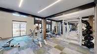 Located at 1033 East Sawmill Road in Flagstaff, Arizona, this two-floor fitness facility offers a comprehensive and thoughtfully designed workout environment spread across two generously sized levels. The space features a contemporary aesthetic defined by bold dark accent beams, warm wood-paneled columns, and multi-toned modular carpet tiles in shades of blue, gray, and green that lend a dynamic energy to the floors.

The lower level is anchored by soaring double-height ceilings that create an open, airy atmosphere. Large windows along the perimeter flood the space with natural light and provide views of the surrounding outdoor environment. A striking motivational mural serves as a focal point, reinforcing the facility's energetic character. This floor houses a robust selection of strength training equipment, including cable machines, weight benches, a power rack, and a well-stocked dumbbell rack, all reflected in wall-to-wall mirrors that enhance the sense of space.

The upper level, connected by a metal-railed mezzanine overlooking the floor below, is dedicated primarily to cardio training. A long row of treadmills, rowing machines, ellipticals, and StairMasters lines the space beneath recessed linear LED lighting. Ceiling fans provide additional comfort throughout. Together, the two floors form a cohesive, well-equipped fitness destination suited for a full range of training needs.