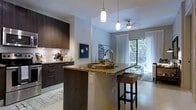 This single-floor residence showcases a thoughtfully designed open-concept layout that seamlessly connects the kitchen and living areas. The kitchen is a standout feature, outfitted with rich dark wood cabinetry that provides generous storage and a striking contrast against the light-toned walls. A granite-topped island serves as the centerpiece, accommodating bar-height seating and functioning equally well as a prep space or casual dining area. Pendant lighting with brushed metal fixtures hangs above the island, adding a refined touch to the space.

The adjoining living area benefits from abundant natural light streaming through a large sliding glass door that leads to a private balcony, offering outdoor views framed by lush greenery. Sheer white curtains soften the light and contribute to the space's airy, contemporary ambiance. A ceiling fan with a built-in light fixture provides both comfort and additional illumination. The light-toned tile flooring runs throughout, tying the kitchen and living zones together cohesively. Overall, the space balances modern aesthetics with functional design in a bright, inviting setting.