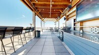 Located at 201 N Water St in the heart of downtown Wichita, this rooftop amenity space offers a well-appointed outdoor experience designed for both relaxation and entertaining. The layout encompasses several distinct zones, each serving a specific function while maintaining a cohesive aesthetic throughout.

At the center of the rooftop is a resort-style pool flanked by a warm wood deck, where lounge chairs are positioned to take full advantage of the sweeping downtown Wichita skyline views. A decorative palm tree adds a distinctive visual element near the pool area.

A substantial covered pergola structure, framed in heavy timber with an open-beam ceiling and strung with Edison-style bulbs, anchors the entertaining zone. Beneath it, a full grilling station with a large gas grill and adjacent bar counter provides ample space for outdoor cooking and serving. Bar-height seating lines one side of the covered area, while a speaker system supports audio throughout.

Adjacent open-air lounge areas feature sectional seating with colorful accent cushions, coffee tables, and large planters, creating comfortable gathering spots. Large-format tile and wood decking cover the expansive floor surfaces throughout. The perimeter railing system ensures unobstructed panoramic views of the surrounding cityscape in all directions.