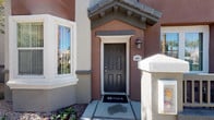 This single-story residence features a welcoming exterior finished in warm terracotta-toned stucco, accented by a decorative gabled entry with a wall-mounted lantern and a classic six-panel door. Manicured landscaping frames the entryway, adding a polished curb appeal.

Inside, the living room is thoughtfully designed with a neutral color palette anchored by warm gray walls and white ceilings. Two plush, cream-upholstered sofas face each other across a rich, dark-wood oval coffee table, creating a comfortable and symmetrical seating arrangement. A bold orange-and-white geometric area rug ties the space together, complementing the vibrant abstract wall art displayed nearby. A dark wood media console along one wall provides practical storage and display space.

Natural light flows generously into the room through multiple windows dressed in coordinating orange Roman shades with vertical blinds, lending a cohesive warmth to the space. Soft carpet underfoot enhances the comfortable, livable atmosphere. The overall ambiance is cheerful and well-appointed, blending contemporary furnishings with warm, earthy accent tones for an inviting everyday living environment.