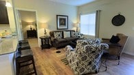 This single-floor space showcases a thoughtfully designed living area that balances comfort and style. The living room features warm hardwood flooring that anchors the space and complements the neutral, light-toned walls. A large window fitted with white horizontal blinds and grommet-top curtains allows natural light to filter in, brightening the room throughout the day.

The seating arrangement includes a plush dark brown sofa adorned with decorative patterned and striped throw pillows, paired with a bold black-and-white floral upholstered armchair that adds a striking visual contrast. A dark wood coffee chest serves as a functional centerpiece, while a round side table with a cross-base holds a classic table lamp. Additional floor lamps on either side of the seating area provide warm ambient lighting.

Wall décor includes a framed botanical print that adds a refined touch to the space. A striped area rug defines the seating zone over the hardwood floor. A glimpse through the doorway reveals an adjacent room, suggesting a connected and practical layout. Overall, the space offers a warm, welcoming ambiance suited for relaxed everyday living.