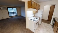 This single-floor residential unit offers a straightforward and functional layout designed for everyday living. The kitchen is efficiently arranged and equipped with a coil-burner electric range, a full-size refrigerator, and ample overhead cabinetry in a warm oak finish. Light-toned vinyl flooring in the kitchen provides easy maintenance, while a built-in under-cabinet light adds task lighting above the range. The kitchen flows seamlessly into the adjacent living area through an open arched passageway, creating a connected feel between the two spaces. The living area features wall-to-wall carpeting in a neutral tone, contributing to a cozy and comfortable atmosphere. A baseboard heater runs along the exterior wall, ensuring efficient warmth throughout the colder months. A large sliding or full-width window at the far end of the living area allows natural light to fill the room and provides a view to a private balcony, which features a metal railing and overlooks a treed outdoor setting. The textured ceiling throughout the unit is a consistent design element, and the overall color palette of warm beiges and browns gives the space a grounded, understated character well-suited for comfortable day-to-day living.