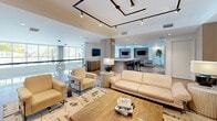 Located at Legacy Gateway in Fort Myers, Florida, this contemporary amenity space spans nearly 2,000 square feet of thoughtfully designed, single-floor communal living. The open-plan layout flows seamlessly between distinct zones, each serving a unique social function while maintaining a cohesive modern aesthetic throughout.

The main lounge area anchors the space with plush cream sofas and armchairs arranged around a bold, terracotta-toned bookshelf wall featuring a centrally mounted flat-screen TV. Geometric track lighting with matte black rails adds an industrial edge to the otherwise warm, neutral palette, while light hardwood flooring runs throughout, tying each zone together.

Adjacent to the lounge, a bar-height counter with sleek black stools serves as a casual gathering and entertaining hub. A dedicated game room houses a full-size billiards table and shuffleboard court, accented by an exposed brick feature wall and floor-to-ceiling windows that invite natural light and outdoor views of palm trees and the resort-style pool.

A standout feature is the private golf simulator room, complete with a projector screen, turf flooring, and club storage — a rare and impressive amenity. An additional seating nook with modern lounge chairs offers a quieter retreat. Elegant pendant chandeliers in the hallway overlook the pool courtyard below, reinforcing the upscale, resort-inspired character of this exceptional amenity space.