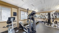 This dedicated fitness center is a compact yet fully equipped space designed to support a comprehensive workout routine. The room features durable rubber flooring throughout, providing a safe and practical surface for exercise. Natural light filters in through multiple windows along the walls, creating a bright and welcoming environment, while fluorescent lighting ensures the space remains well-lit at all times. A large wall-mounted mirror runs along one side of the room, visually expanding the space and allowing users to monitor their form during workouts. The cardio section is outfitted with a treadmill, an elliptical trainer, and a stationary bike, offering a variety of aerobic exercise options. A multi-station cable weight machine provides a full range of strength training exercises, complemented by a rack of dumbbells in varying weights for free weight workouts. A wall-mounted television is positioned for easy viewing during cardio sessions. The space is accessed through a set of classic French doors, which contribute a touch of architectural charm. A ceiling fan aids in air circulation, maintaining comfort during intense workouts. The warm beige walls and wood-paneled ceiling add a cozy, inviting character to the fitness environment.