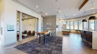 Located at 8025 Ohio Dr in Plano, TX, this thoughtfully designed single-floor community clubhouse offers a generously proportioned gathering space finished with refined, contemporary details throughout. The open layout is anchored by elegant herringbone-patterned tile flooring, crisp white walls, and decorative columns that define distinct social zones without interrupting the natural flow of the space.

A central lounge and meeting area features a striking live-edge wood communal table surrounded by modern chairs, set atop a textured area rug. Adjacent to it, a billiards zone showcases a full-size pool table beneath a sculptural multi-pendant brass and matte black light fixture, with dual wall-mounted flat-screen televisions nearby for entertainment.

A curved sunroom alcove lined with tall, floor-to-ceiling windows floods the space with natural light and offers sweeping outdoor views of the surrounding landscape, furnished with high-top bistro tables and leather bar stools — ideal for casual gatherings.

The fully equipped kitchen features rich dark wood cabinetry, expansive white quartz countertops, stainless steel appliances including a full-size refrigerator, and dramatic copper pendant lighting. A quieter lounge nook with soft seating provides a comfortable retreat. Together, these spaces create a sophisticated, versatile environment well-suited for both social events and everyday community use.