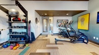 This fitness center is designed to support a comprehensive range of workout routines within a single, open floor. The space features a distinctive patterned hardwood-style floor with interlocking light and teal-toned planks arranged in a geometric layout, adding visual interest underfoot. The ceiling incorporates a recessed dark-paneled tray detail with embedded recessed lighting, complemented by a ceiling fan that promotes air circulation throughout the room.

Cardio equipment is well-represented, with multiple treadmills, ellipticals, and a stationary bike positioned to make use of both wall-mounted televisions and natural light from roller shade-equipped windows. A full-length mirrored wall with a ballet barre runs along one side of the room, making the space well-suited for stretching, yoga, or barre workouts. A multi-functional cable and strength machine is positioned nearby, along with a adjustable weight bench.

On the opposite side, a dedicated storage rack holds an assortment of free weights, resistance bands, medicine balls, and kettlebells. Colorful original artwork is displayed throughout, adding a vibrant, gallery-like quality to the space. A glass-door entryway connects to an adjoining room, further extending the overall functionality of the fitness facility.